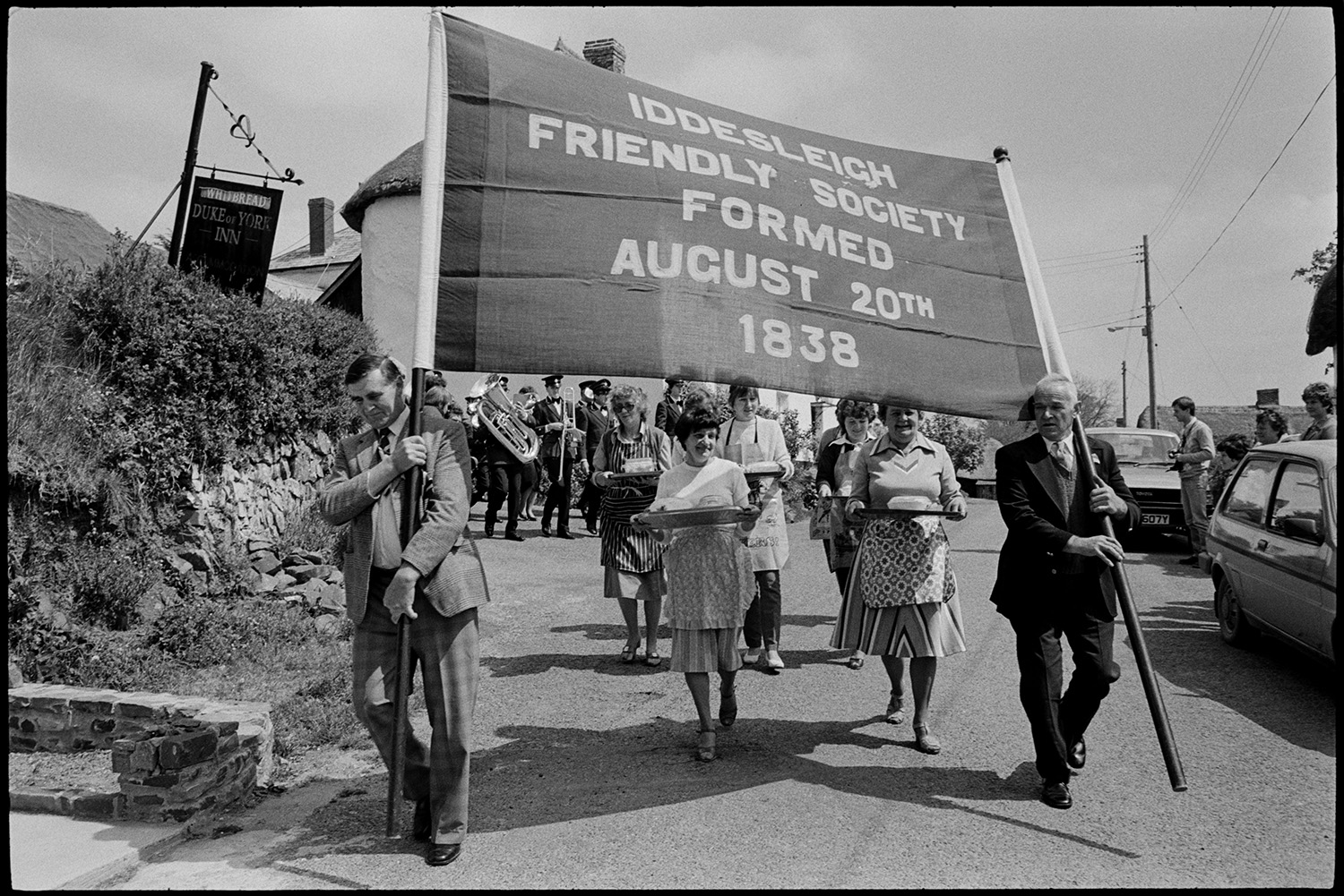 Club Day, women and club members parading down to village hall with mashed potatoes.
[Women wearing aprons and carrying trays with bowls of mashed potato along a street in the Iddesleigh Club Day parade. They are walking behind the Iddesleigh Friendly Society banner with the Hatherleigh Silver Band following them.]