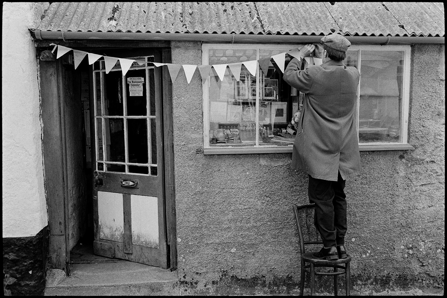Club Day, man decorating shop with bunting, flags.
[Tim Laing standing on a chair decorating a shopfront with bunting for Club Day at Iddesleigh. Greetings cards are displayed in the shop front window and the shop roof is made from corrugated iron.]