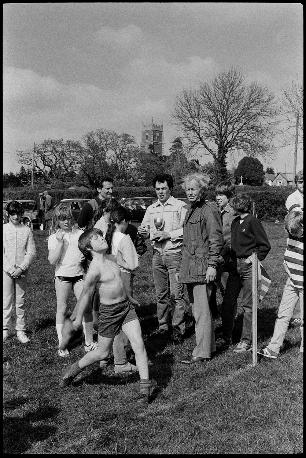 Club Day. Sports, running races, wellie throwing, prizes being given out.
[Children and adults gathered in a field for sports at Iddesleigh Club Day. A boy is about to throw a wellie. Iddesleigh church tower is visible in the background.]