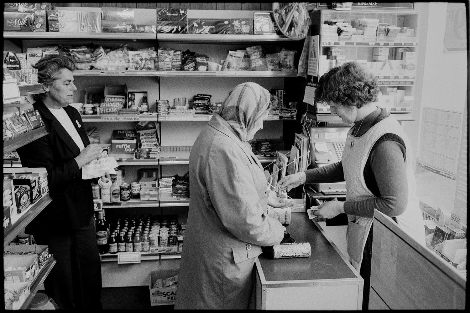 Shopkeepers in village store, customers, women buying at till.
[Women queuing up and paying for goods at the till in the village shop at Winkleigh. Sweets, soft drinks and cigarettes are on display on the shelves.]
