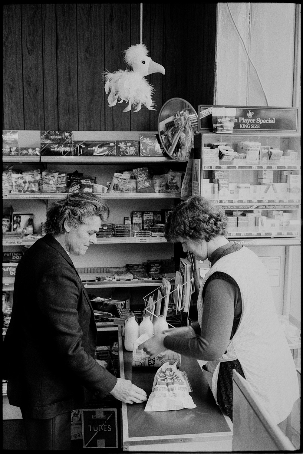 Shopkeepers in village store, customers, women buying at till.
[A woman paying for goods at the till in the village shop at Winkleigh. Sweets, soft drinks and cigarettes are on display on shelves. A fluffy bird is hanging from the ceiling and milk bottles are on the shop counter.]