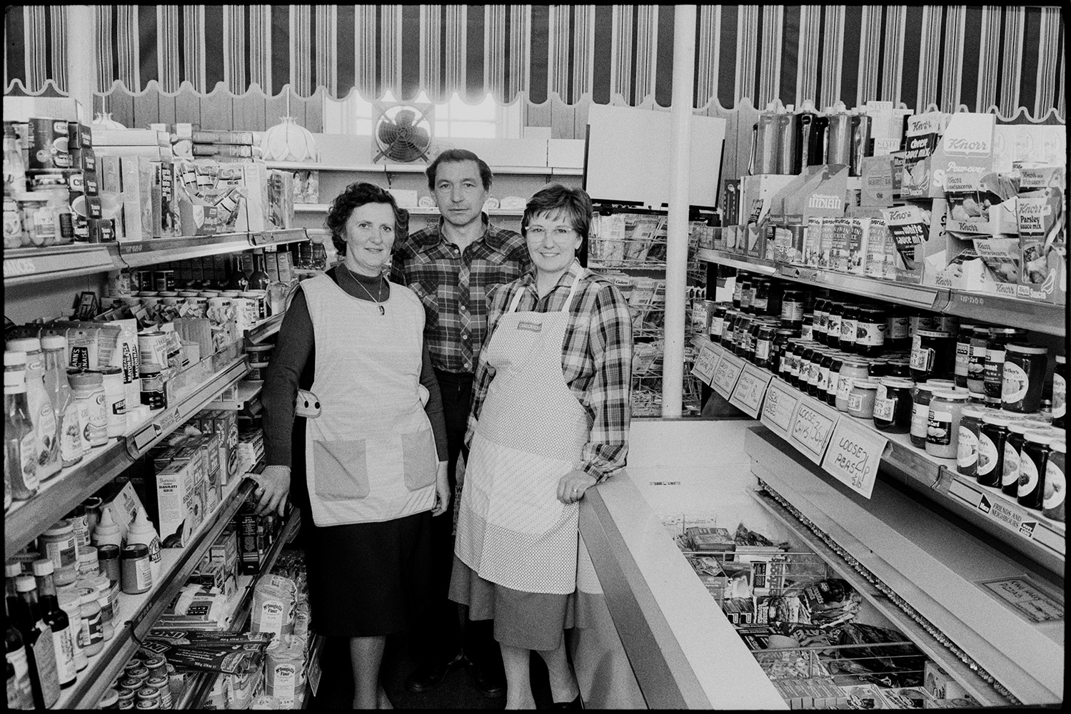 Shopkeepers in village store, customers, women buying at till.
[Three shopkeepers in the village shop at Winkleigh standing in an aisle with various jars and packets of food displayed on shelves and in a freezer cabinet.]