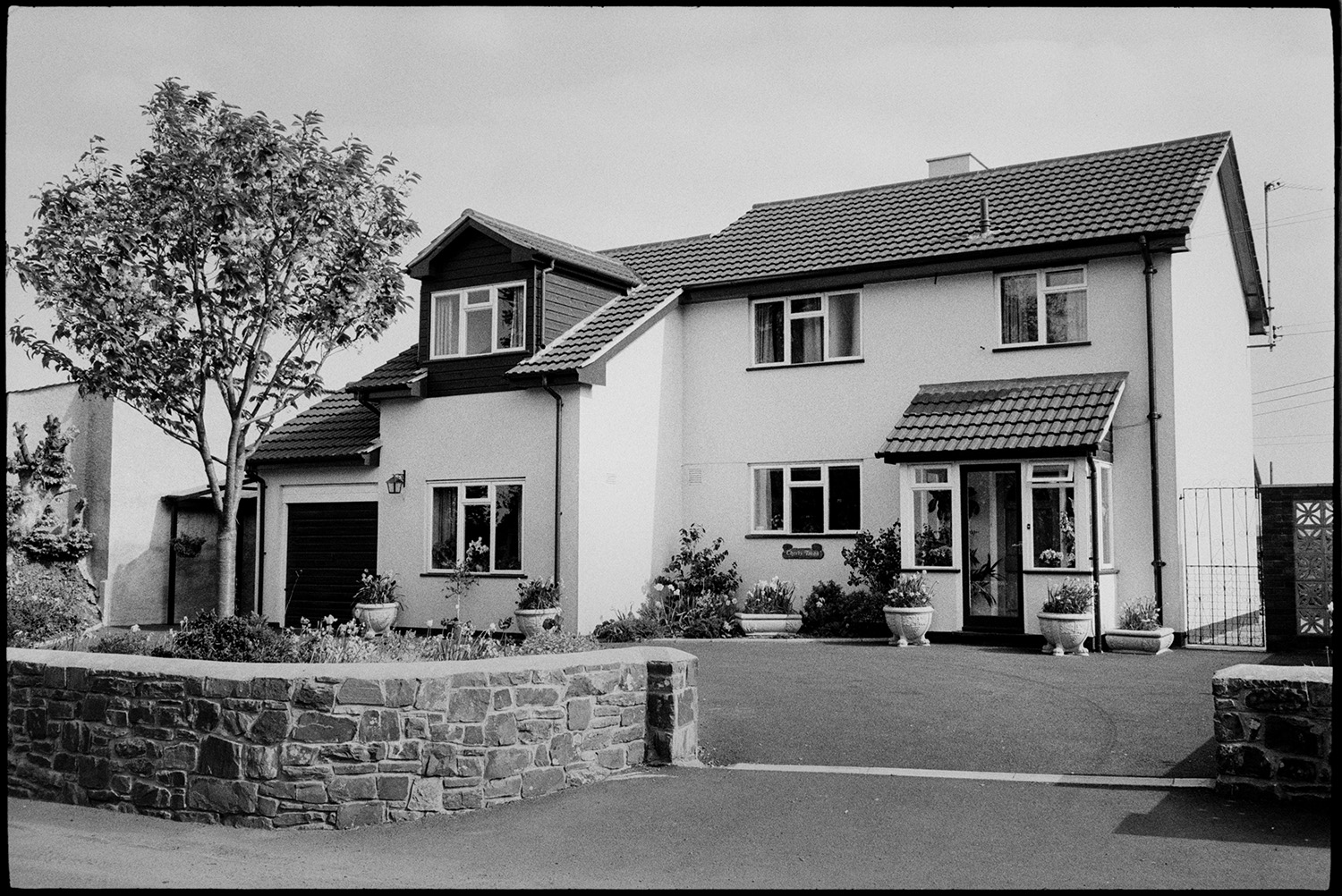 Modern house in village, example of hideous new housing.
[A modern house in Kings Nympton with a garage and tiled roof. Flower pots are in front of the house on the driveway.]