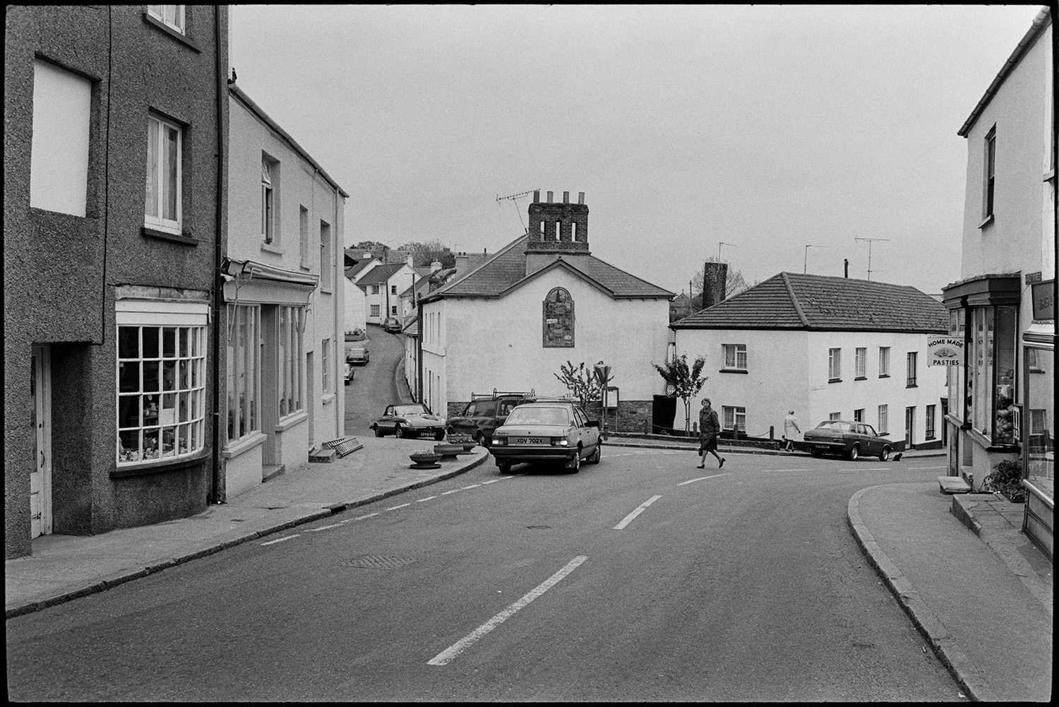 Street scene with pub.
[A view of a street in Hatherleigh with a pasty shop, parked cars and a woman crossing the road.]