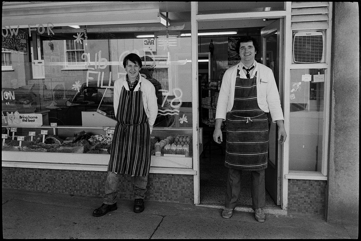Two butchers in front of shop.
[Two butchers wearing striped aprons standing outside their butcher's shop in North Tawton. Meat is displayed in the shop front window.]
