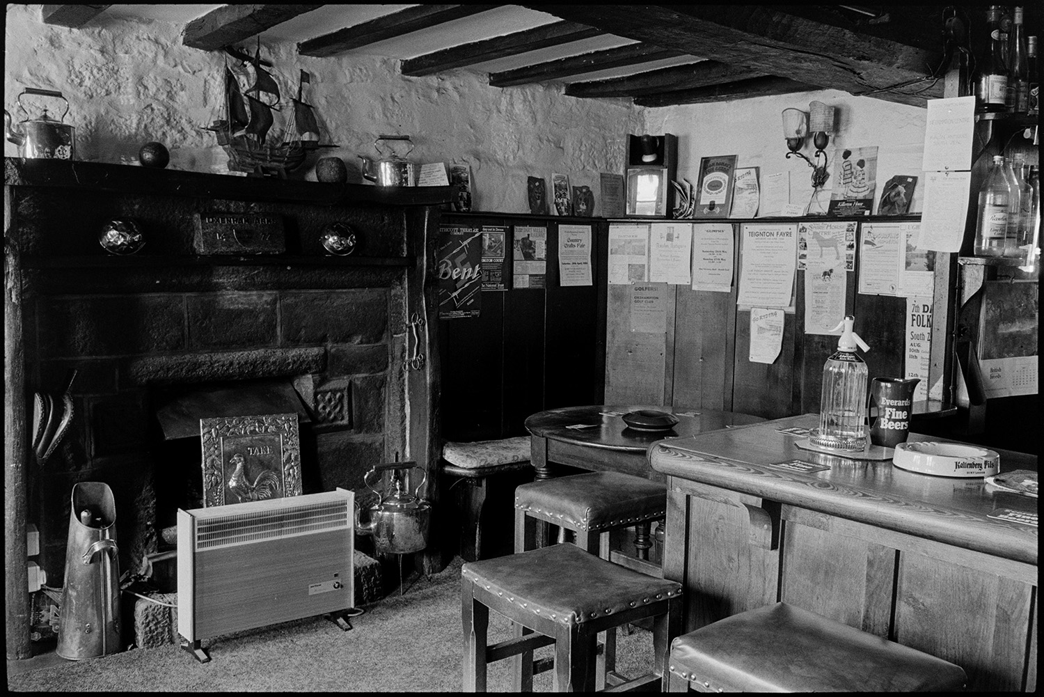 Woman ironing in pub sitting room with Television, fireplace, pub bar.
[A fireplace with electric heater and corner of the bar at the Kings Arms, South Zeal. Posters are displayed in a settle next to the bar and various ornaments are around the fireplace, including kettles and the model of a ship.]