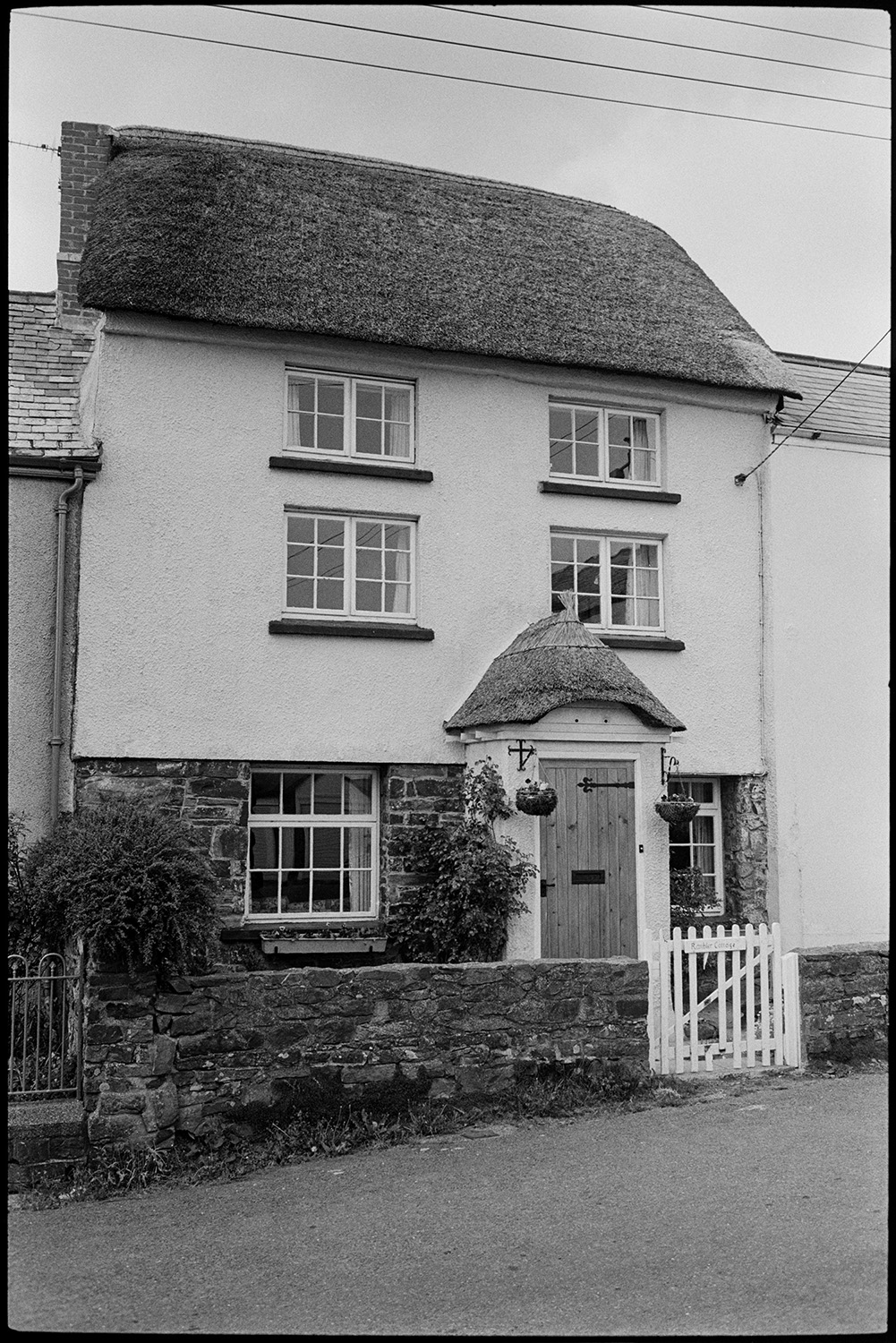 Modernised thatched cottage example of hideous conversion, see old photo.
[The front of Rambled Cottage at High Bickington. The three storey thatched cottage has been modernised. Hanging baskets decorate the door in the thatched porch. The gate ]