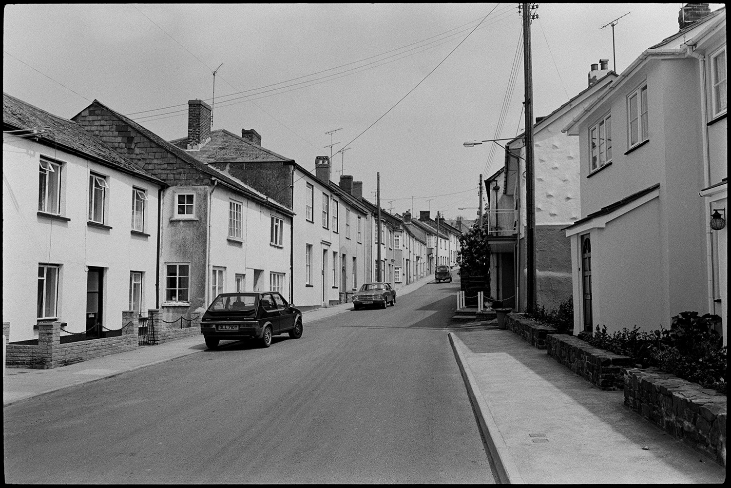 Comparison with old photo. Street scenes, man mowing lawn. Lorries and cars.
[A street at North Tawton with cottages, houses and parked cars.]