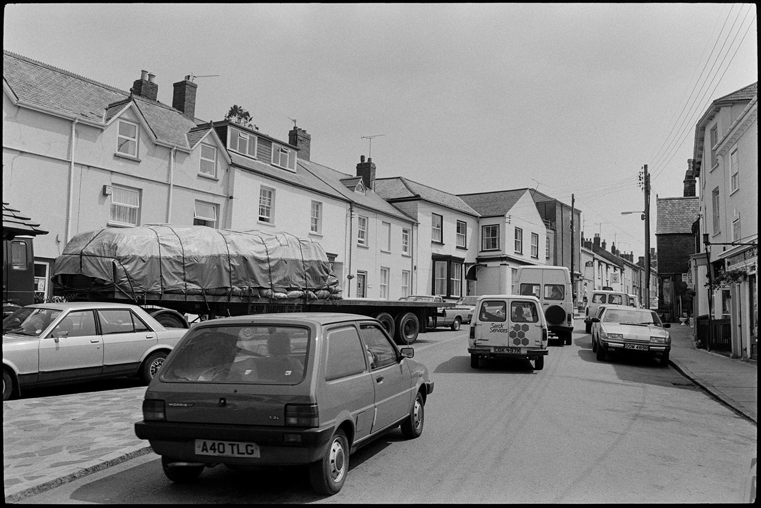 Comparison with old photo. Street scenes. Man mowing lawn. Lorries and cars.
[A busy street in North Tawton with houses, cars, vans and a lorry.]