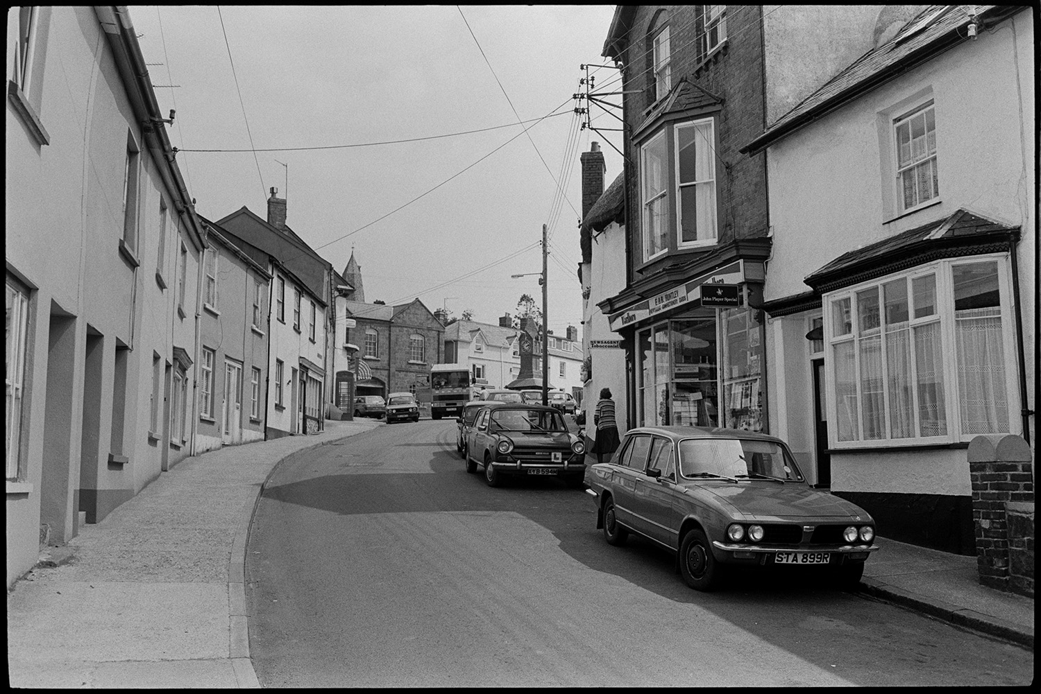 Comparison with old photo. Street scenes. Man mowing lawn. Lorries and cars.
[A street in North Tawton, looking towards the clock tower. Cars are parked outside E & B Huntley, newsagents shop front.]
