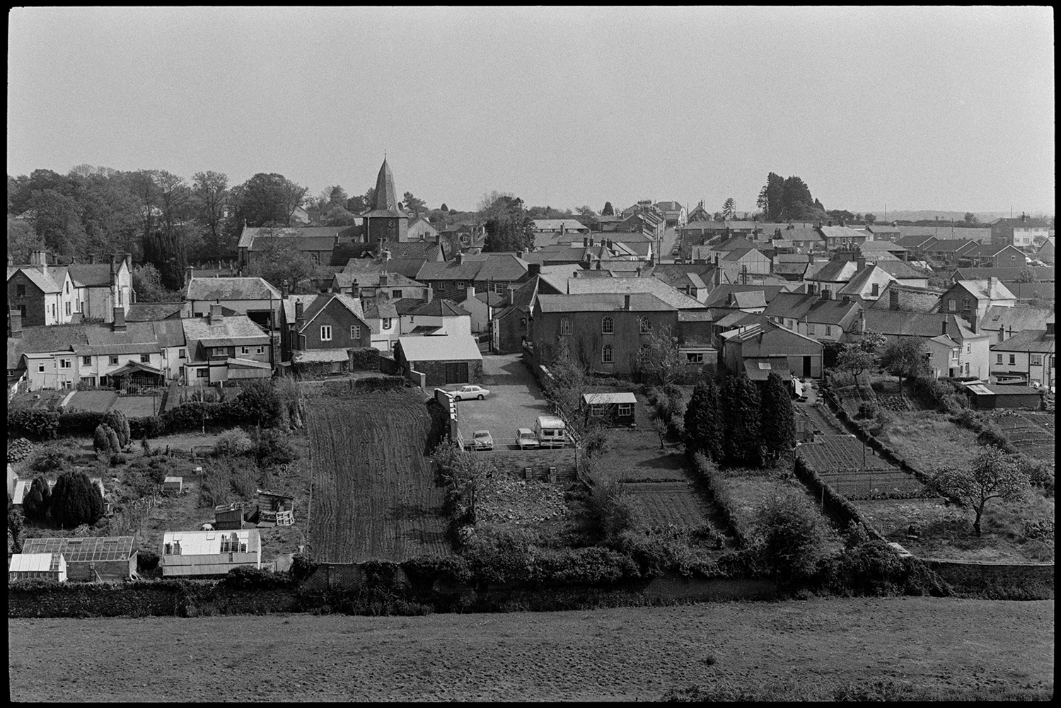 Comparison with old photo. Man gardening with town in background.
[A view of North Tawton taken from a hillside showing the town buildings, gardens, greenhouses and church.]