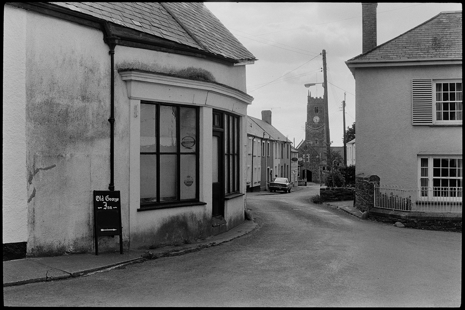 Former village shop and street, comparison with old photo.
[A view looking down the street from the former shop to the church tower in High Bickington. A sign points to the Old George Inn and parts of Howards House can be seen opposite the shop.]