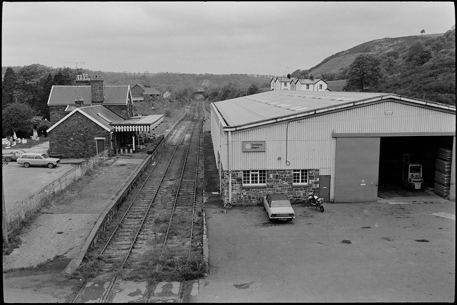 Scenes around town, comparison with old photos, railway line and station, lorries and cars, river.
[A scene taken from a bridge over the railway station and railway lines running past the ICI fertilizer depot, at Torrington. The platform, station buildings and parked cars are visible.]