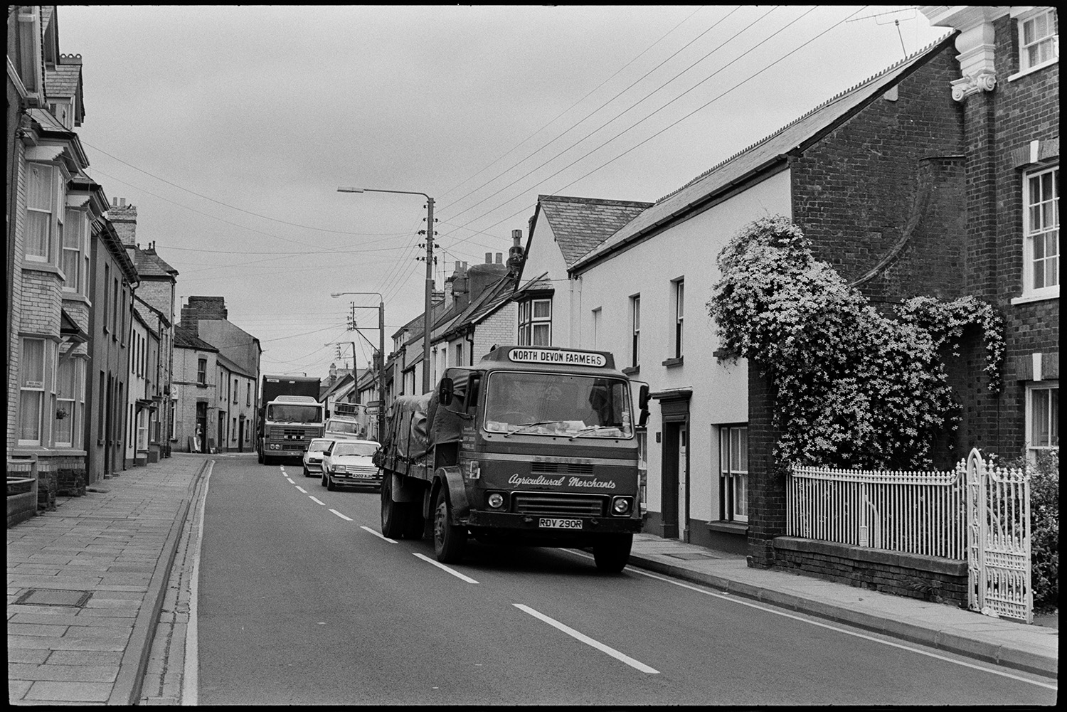 Scenes around town, comparison with old photos, railway line and station, lorries and cars, river.
[Lorries and cars driving through a street in Torrington. A North Devon Farmers lorry is in the foreground next to a building with railings and clematis growing up it.]