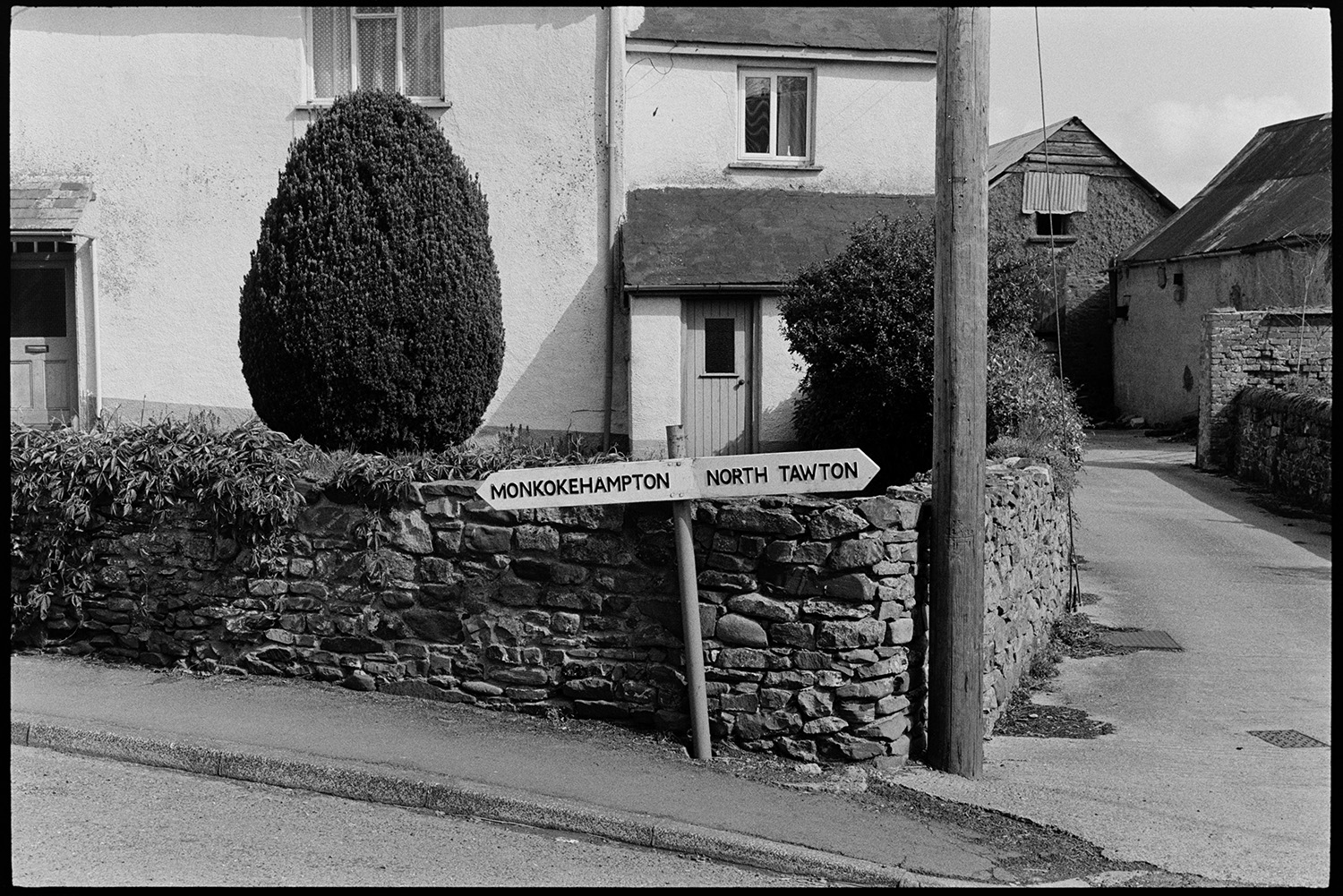 Road sign on corner of farm wall, new sign replacing nicer old one, see earlier pic?
[A new signpost at Exbourne by a farm garden wall pointing the way to Monkokehampton and North Tawton. Barns can be seen in the background.]