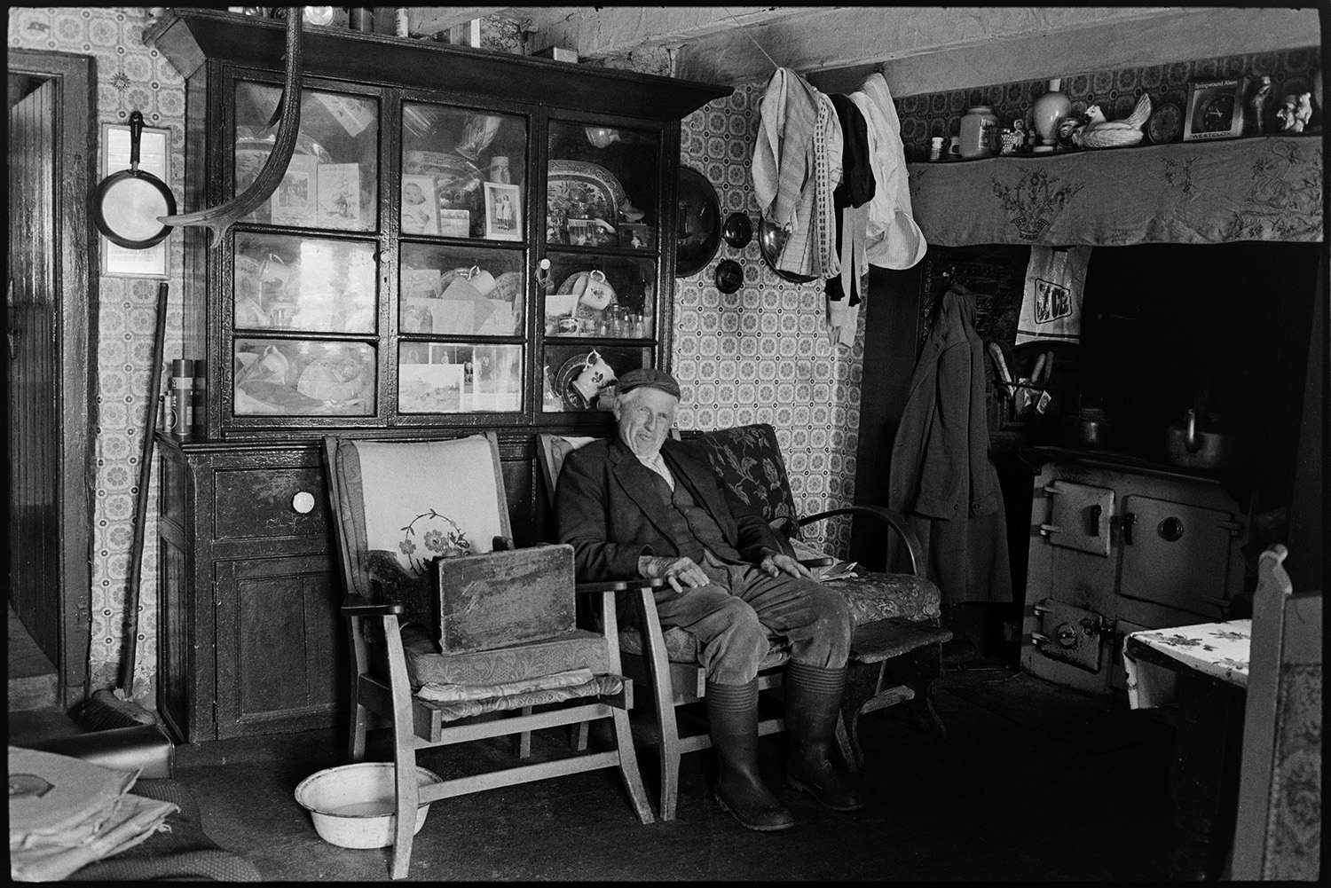 Farm interior, man seated with dresser, furniture. Now modernised.
[Wilfie Spiers sitting near the Rayburn at Mount Pleasant, Beamsworthy. A dresser, ornaments on the mantelpiece and coats and washing hanging up to dry are visible in the room. The walls are covered with patterned wallpaper.]