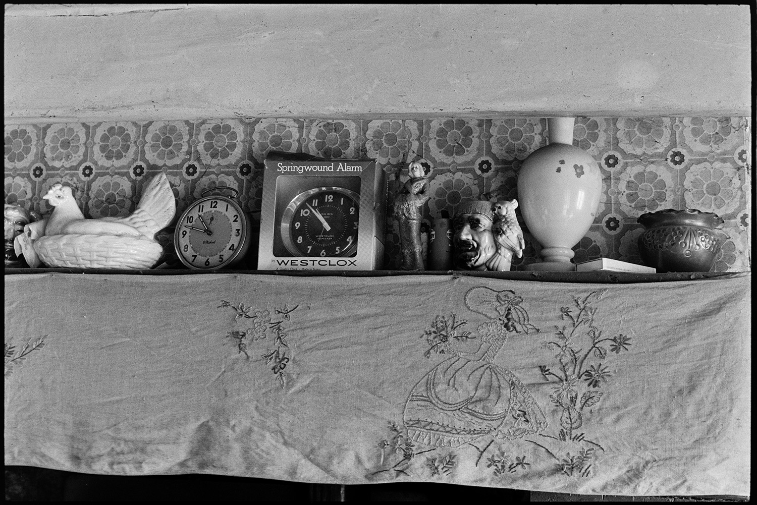 Clock and ornaments on farmhouse mantelpiece, embroidered cloth.
[Clocks, a vase and ornaments on a mantelpiece trimmed with a hand embroidered cloth at Mount Pleasant, Beamsworthy. One of the clocks is in its packaging.]