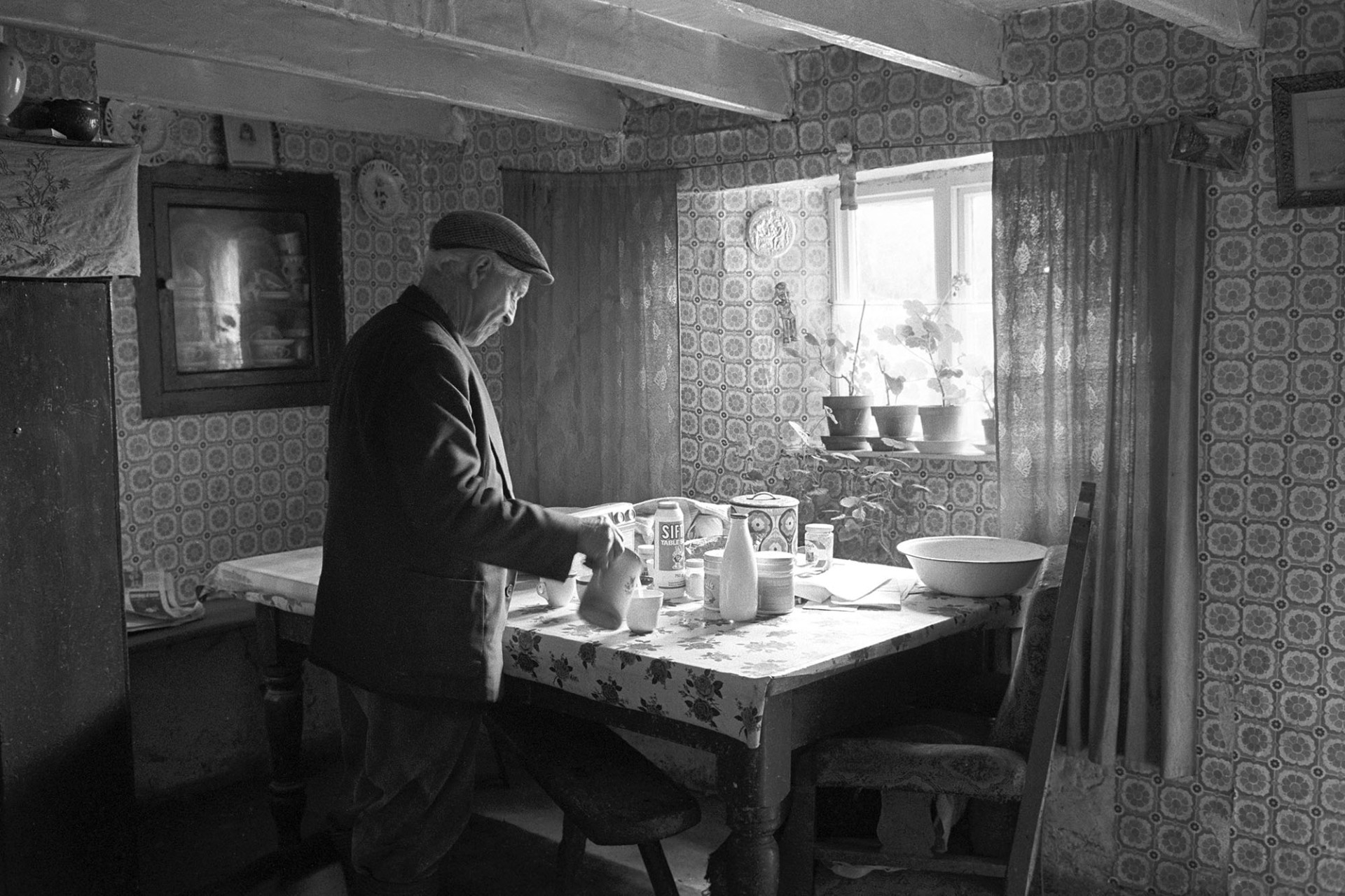 Man pouring cup of tea at farmhouse table, pot plants in window. 
[Wilfie Spiers making a cup of tea on the table in his kitchen at Mount Pleasant, Beamsworthy. The walls are covered in patterned wallpaper ad pot plants are in the windowsill. Beams are also visible on the ceiling.]