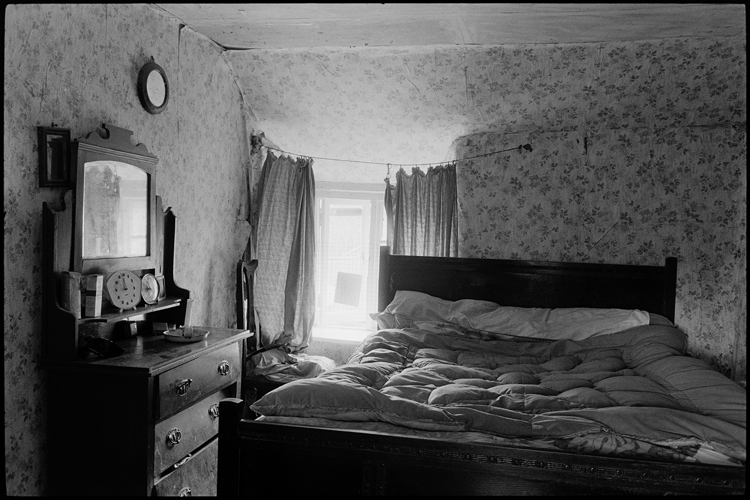 Farmhouse bedroom with clocks dressing table with mirror curtains.
[A farmhouse bedroom at Mount Pleasant, Beamsworthy.  The room contains a bed with a quilt and a dressing table with two clocks and a mirror.  The half drawn curtains are hanging from a wire line.]