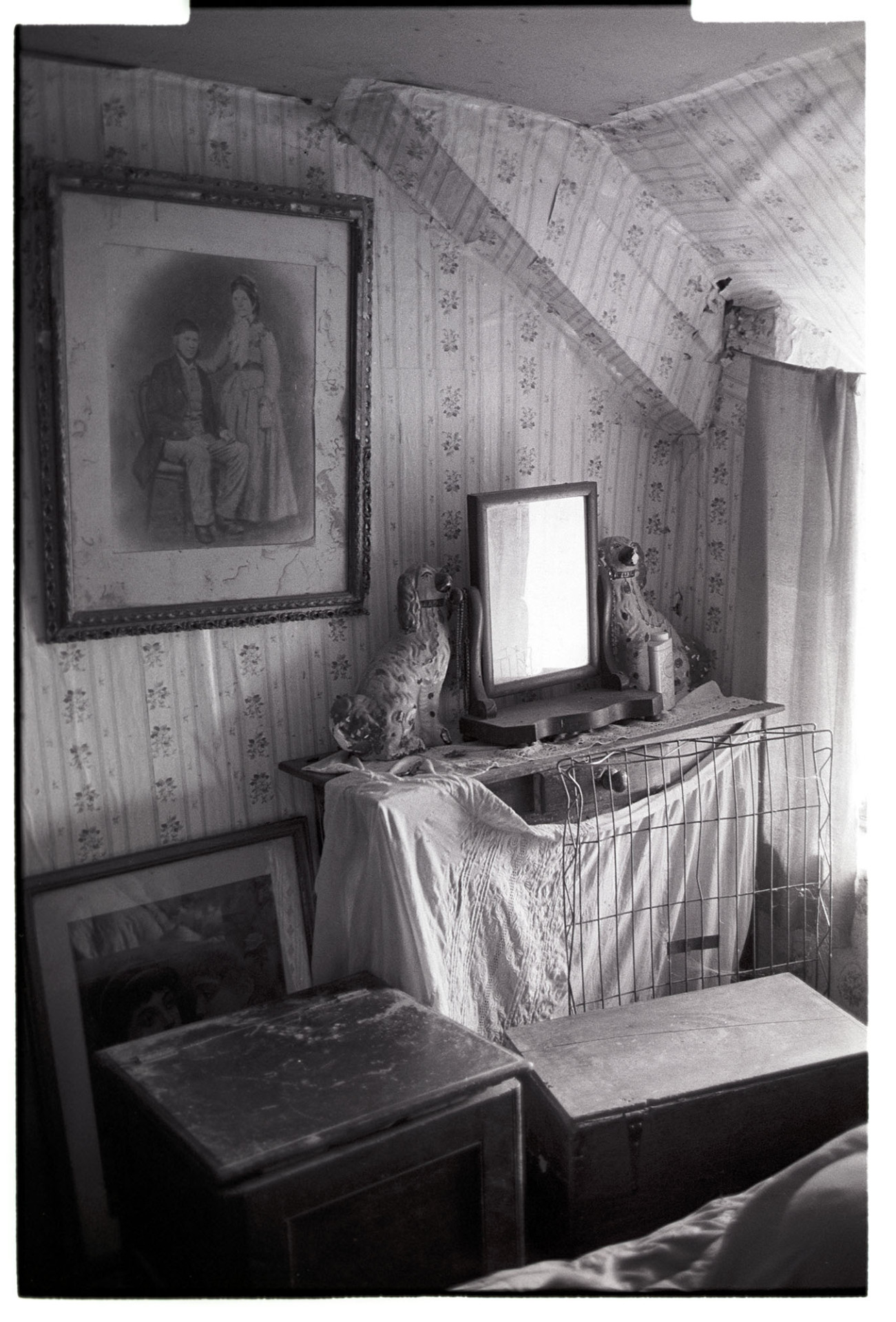 Disused farmhouse bedroom, dressing table with mirror, photograph and china dogs. 
[A disused bedroom in Wilfie Spiers's farmhouse at Mount Pleasant, Beamsworthy. A dressing table with mirror and two china dogs, cupboards and paintings are in the room. The walls are covered with patterned wallpaper.]