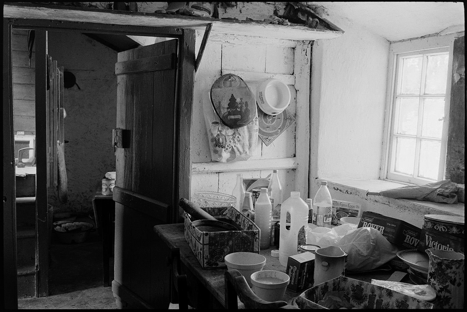 Farmer standing in kitchen, utensils etc, window.
[Utensils, bottles and boxes on a table next to a window and open wooden door at Mount Pleasant, Beamsworthy.]