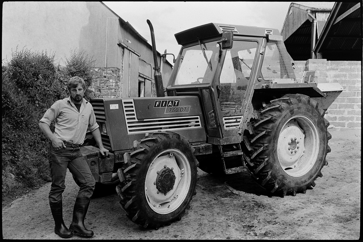 Farmer standing with tractor.
[Simon Berry standing next to a Fiat tractor in a farmyard at Harepath, Beaford. Barns are visible in the background.]