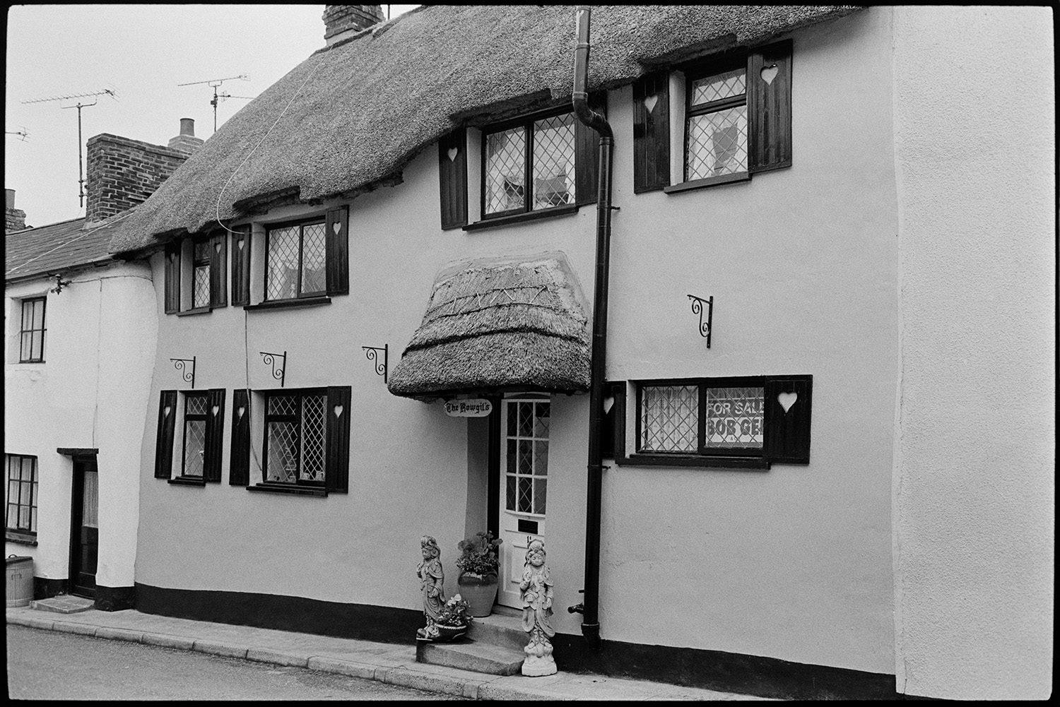 Street with hideously ruined thatched cottage, unsuitable shutters and windows. Aaaaagh!!!
[A thatched cottage with shutters and ornaments on the doorstep for sale in Hatherleigh. Heart shapes are cut into the shutters and a sign below the porch reads 'The Rowgil's'.]