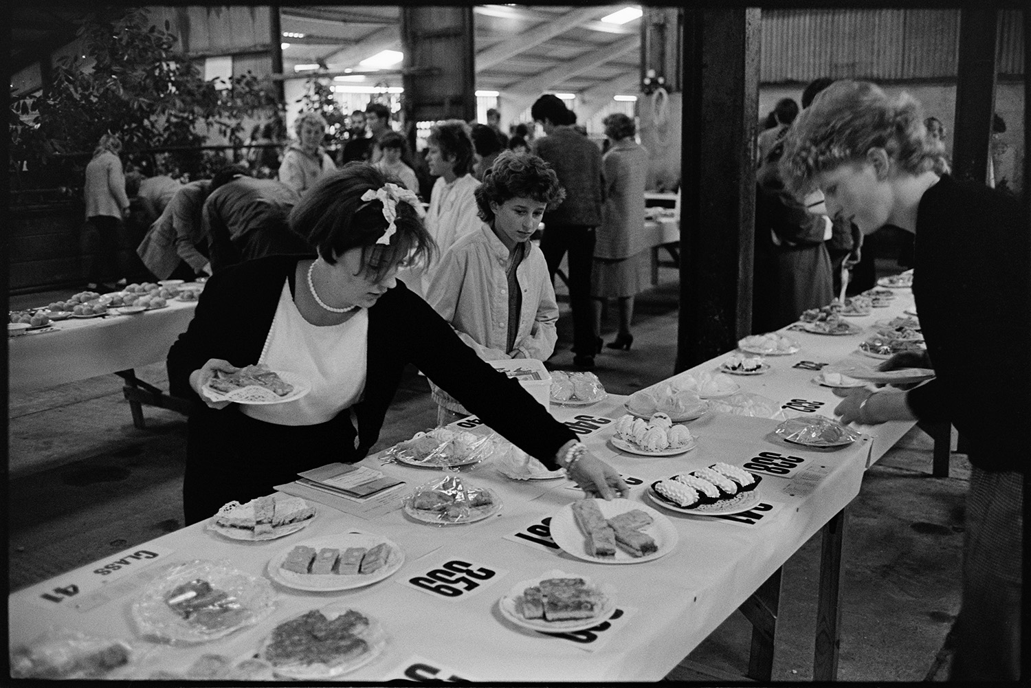 Produce at show, cakes and competitions.
[Women laying cakes out for a competition at Hatherleigh Show. People looking at other produce can be seen in the background.]