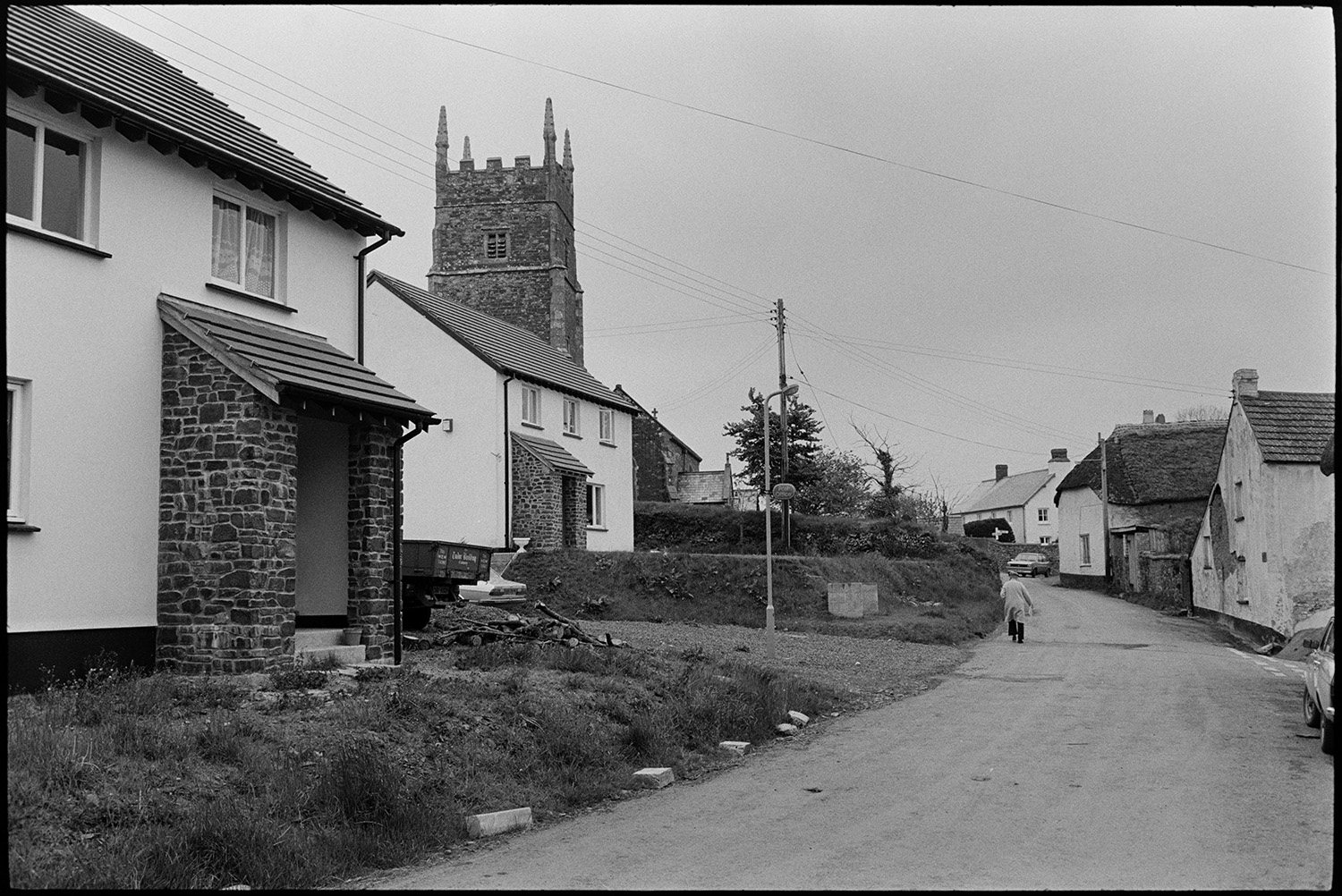 Village with church tower, comparison with old photo.
[ A man walking up a street past cottages, towards the church in Roborough. Two new houses can be seen on the left. ]