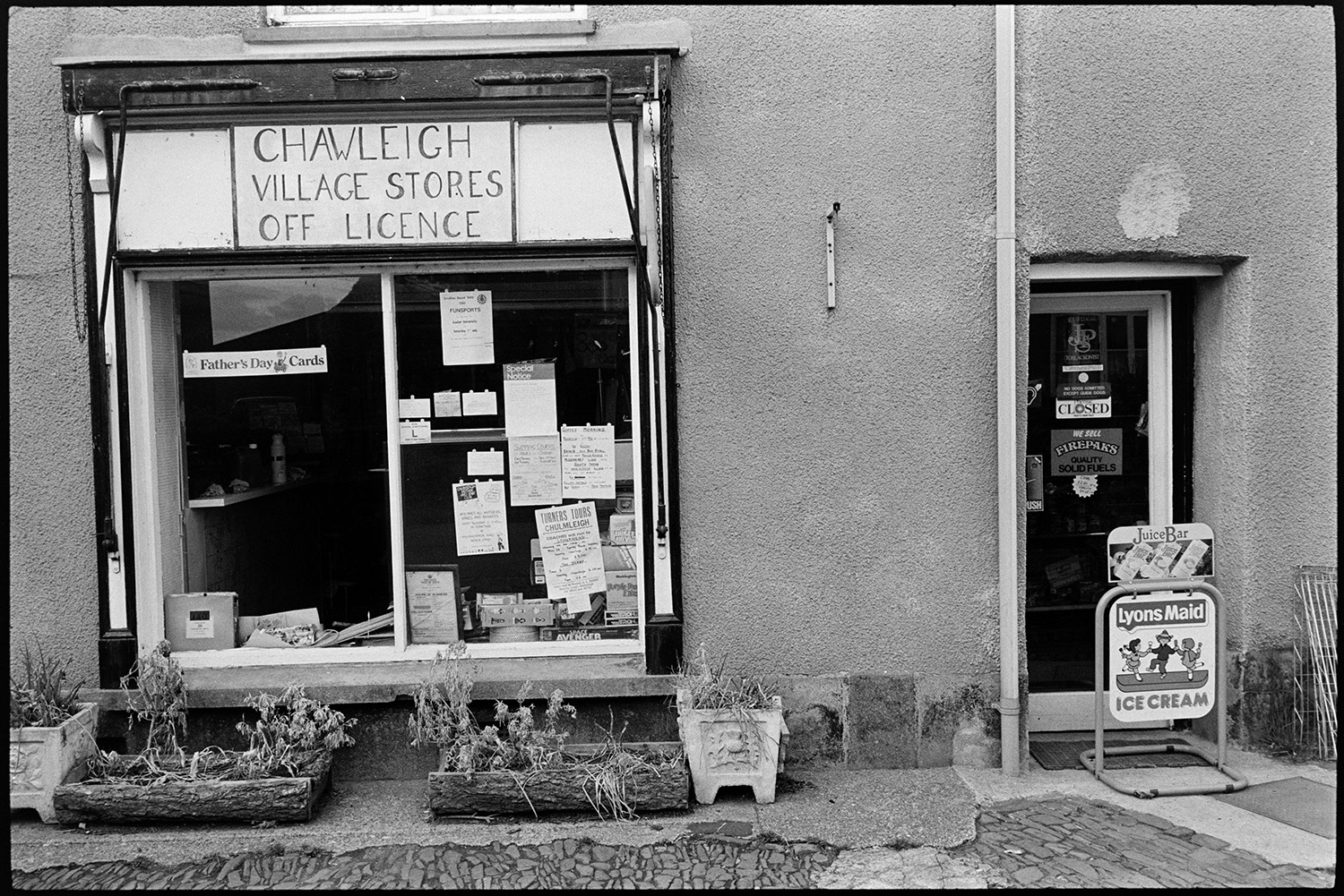 Front of village store and off licence.
[The shop front window of Chawleigh Village Stores and Off Licence. Flower boxes are outside and signs and notices are stuck in the window. The shop is closed.]