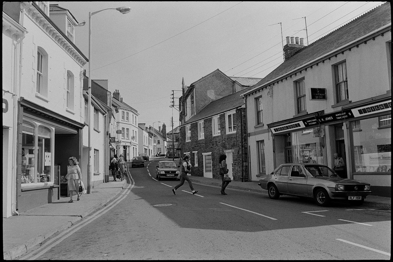 Street with bicycle and cars comparison with old photo.
[A view of a street in Northam showing pedestrians, cars and shops, including  E. R. Jones newsagents.]