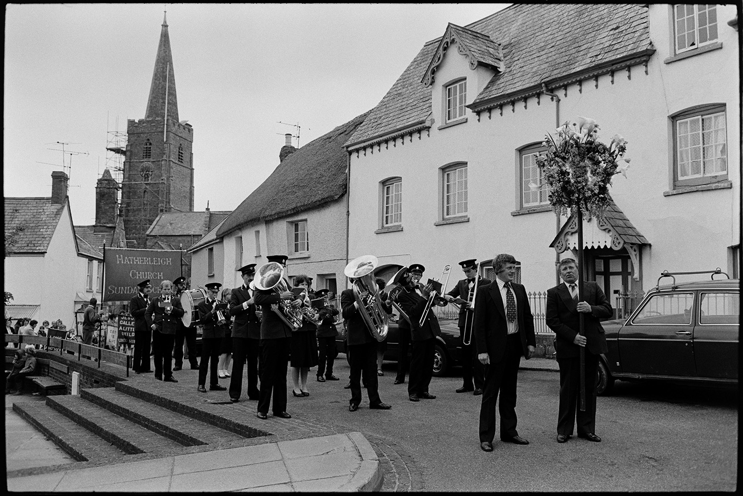 Church Sunday School Parade with banners and Silver Band setting off.
[The Hatherleigh Silver Band preparing to lead the Hatherleigh Church Sunday Schools Parade, with two men in front of the band carrying a floral garland. Behind the band a large banner is being carried and the tower of the church of St John the Baptist and a building with a thatched roof can be seen in the background.]