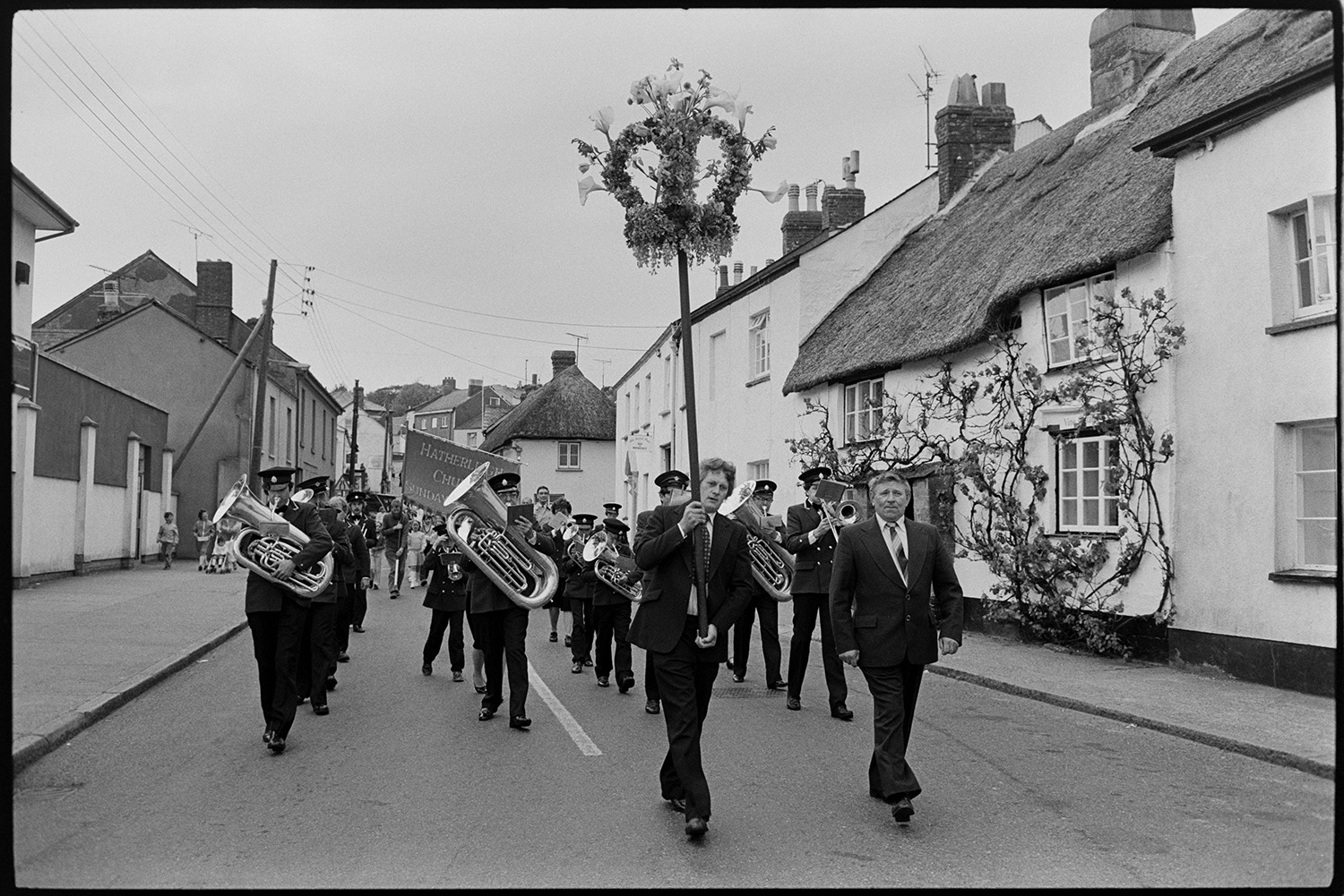 Church Sunday School Parade with banners and Silver Band marching behind garland.<br />
[The Hatherleigh Silver Band leading the Hatherleigh Church Sunday School Parade along a street in Hatherleigh, with two men in front of the band holding a floral garland. Behind the band a large banner is being carried. They are passing a thatched cottage with a rose growing up the front.]