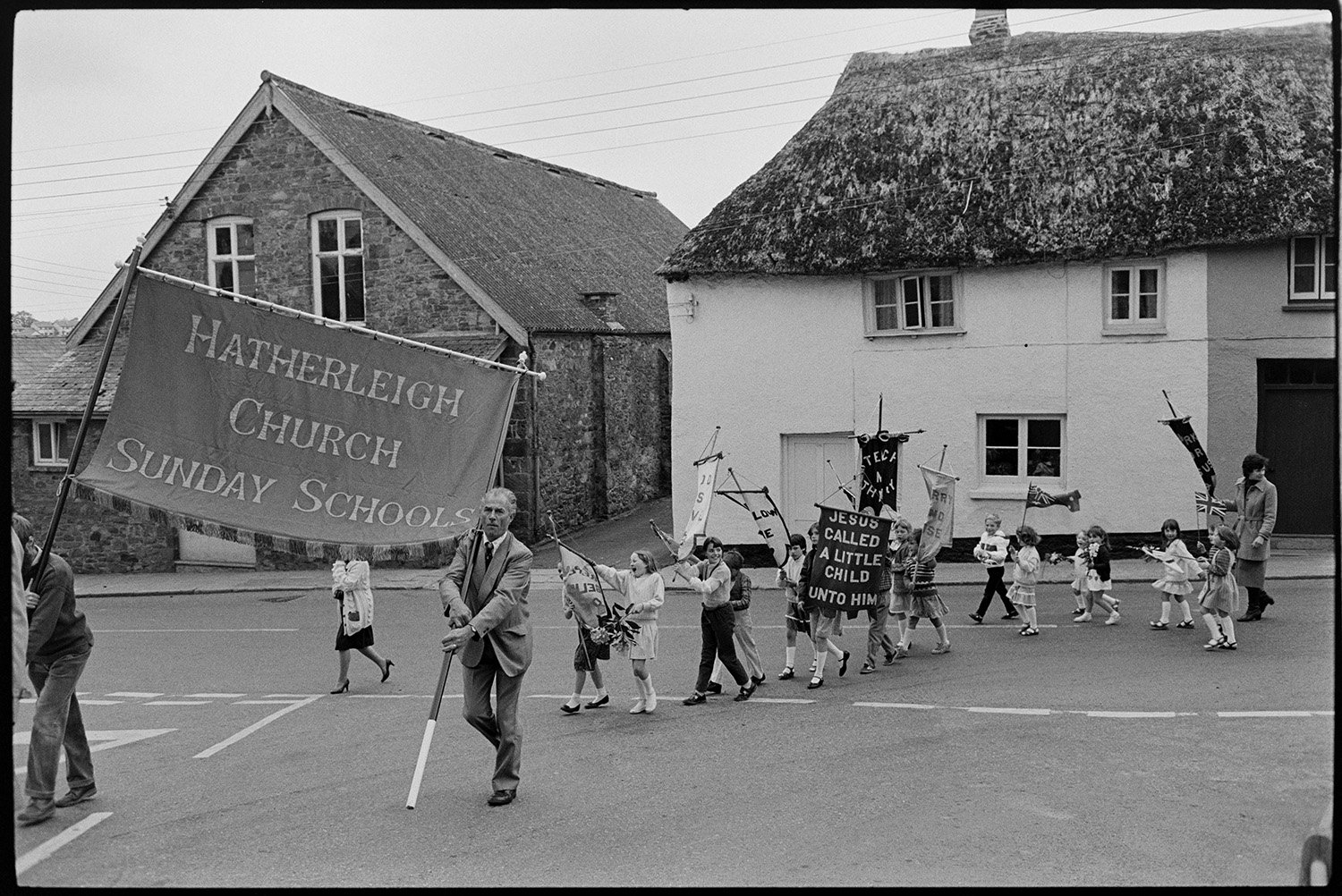 Church Sunday School Parade with banners and Silver Band marching behind garland.
[A line of children walking in the Hatherleigh Church Sunday Schools Parade, being led by two men carrying a large banner. The children are carrying smaller banners. Behind the children is a house with a thatched roof.]