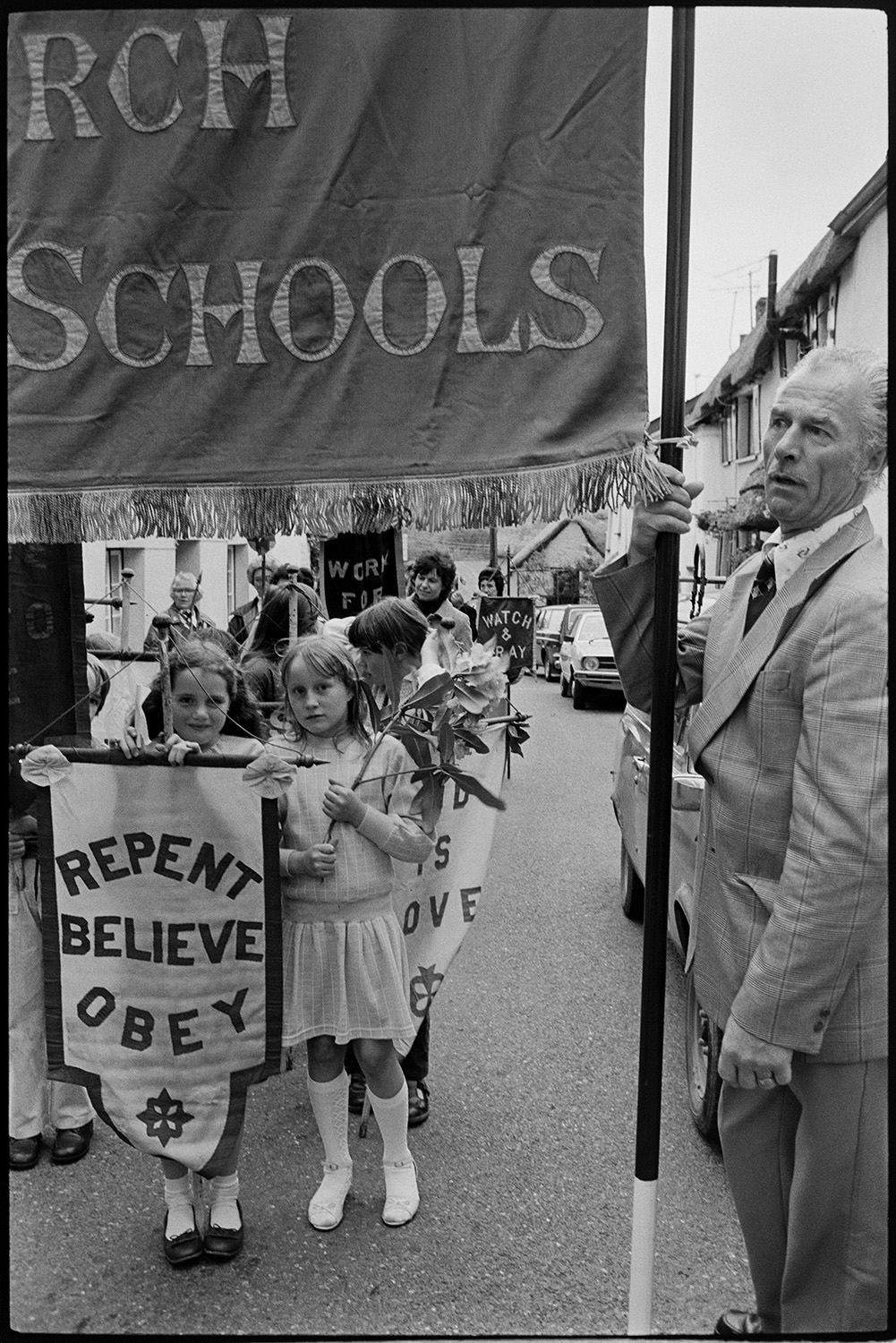 Church Sunday School Parade with banners and Silver Band marching behind garland.
[Children in the Hatherleigh Church Sunday Schools Parade standing in a road in Hatherleigh behind a man carrying a large banner. The children are carrying smaller banners.]