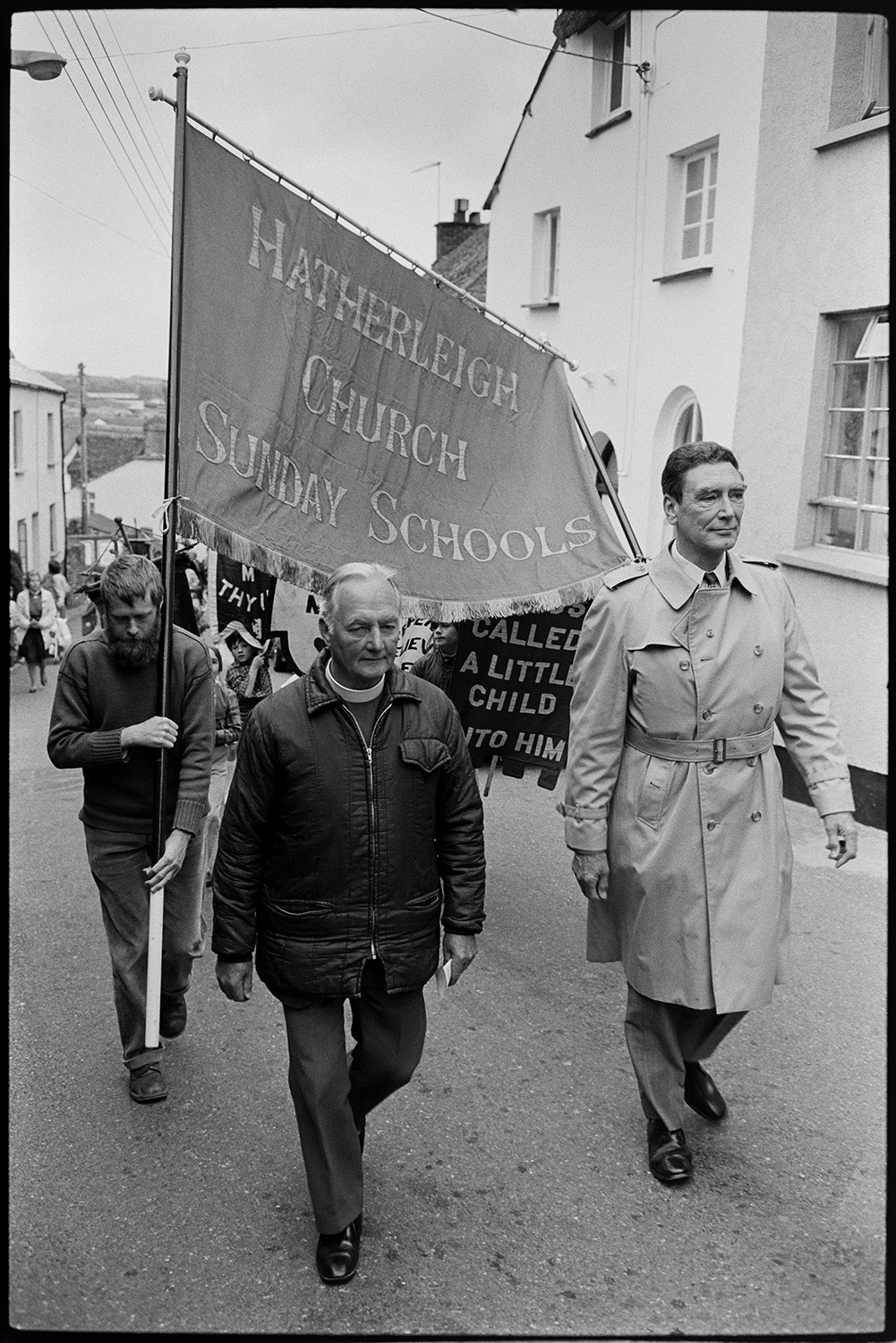 Church Sunday School Parade with banners and Silver Band marching behind garland.
[Vicar and a man marching along a street in Hatherleigh in the Hatherleigh Church Sunday Schools Parade, with a large banner being carried behind them. Children are carrying smaller banners in the background.]