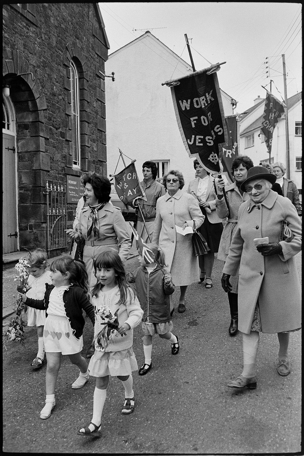 Church Sunday School Parade with banners and Silver Band marching behind garland.
[Women and children marching along a street in Hatherleigh in the Hatherleigh Church Sunday Schools Parade. The children are carrying bouquets of flowers and some of the women are carrying banners.]