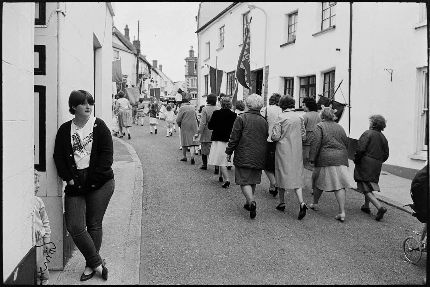 Church Sunday School Parade with banners and Silver Band marching behind garland.
[Women and children marching along a street in Hatherleigh in the Hatherleigh Church Sunday Schools Parade, with some of the women carrying banners. A woman and child are standing in front of a doorway in the foreground, watching the parade.]