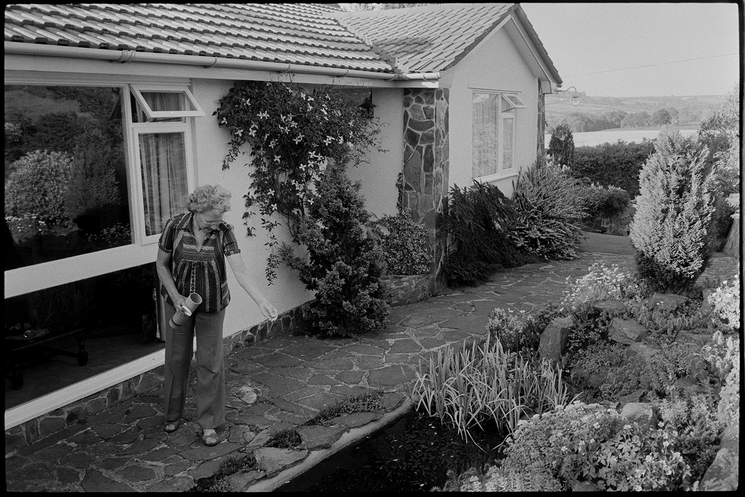 Woman sitting in garden of bungalow.
[Beatrice Nancekivel, also known as Beat, in front of her bungalow at The Patch, Woodtown, Dolton. She is spreading feed on the fish pond.]