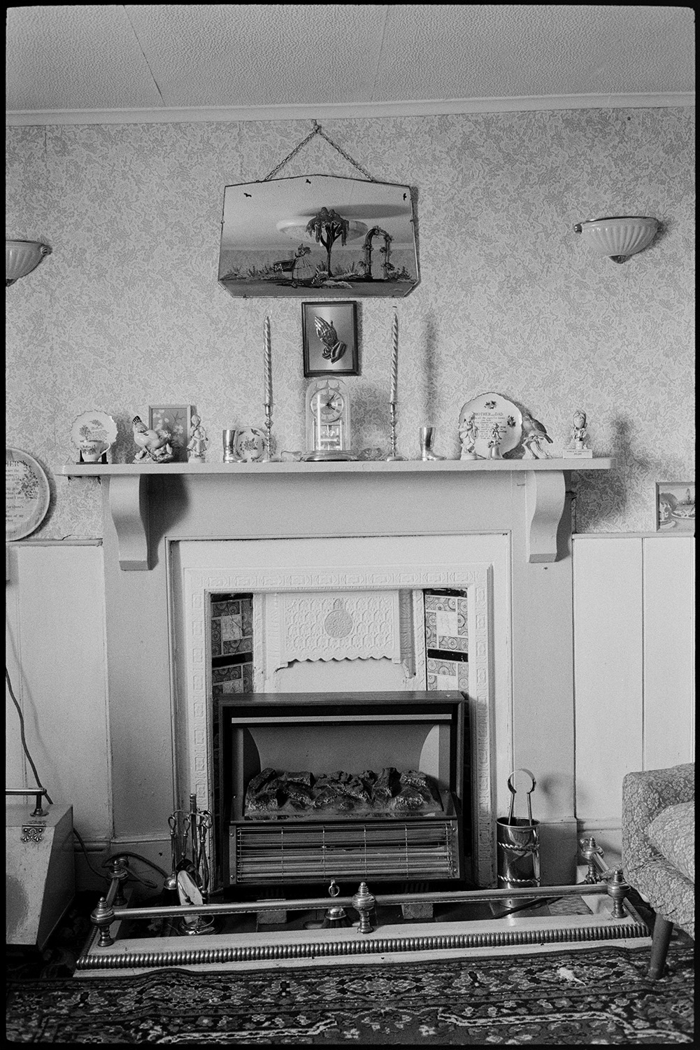 Interior mantelpiece, mirror, comparison with old photo. Ashton, tailor's house.
[Interior of a house in Tricks Terrace, Beaford, which used to belong to Mr Ashton, showing a fireplace with electric fire and mantelpiece. Ornaments and a clock are displayed on the mantelpiece and two pictures are hung on the wall. Mr Ashton was a tailor. An earlier image of the house decorated after his death can be found in the Beaford Old Archive at b00848.]