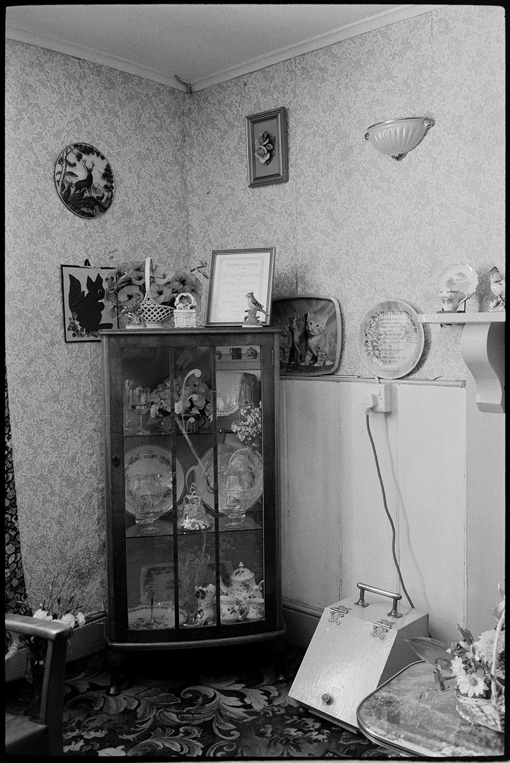 Interior mantelpiece, mirror, comparison with old photo. Ashton, tailor's house.
[Interior of a house in Tricks Terrace, Beaford, which used to belong to Mr Ashton, showing a display cabinet containing china pieces. Four pictures and a plate are hung on the wall nearby, which is covered with patterned wallpaper. Mr Ashton was a tailor. An earlier image of the house decorated after his death can be found in the Beaford Old Archive at b00848.]