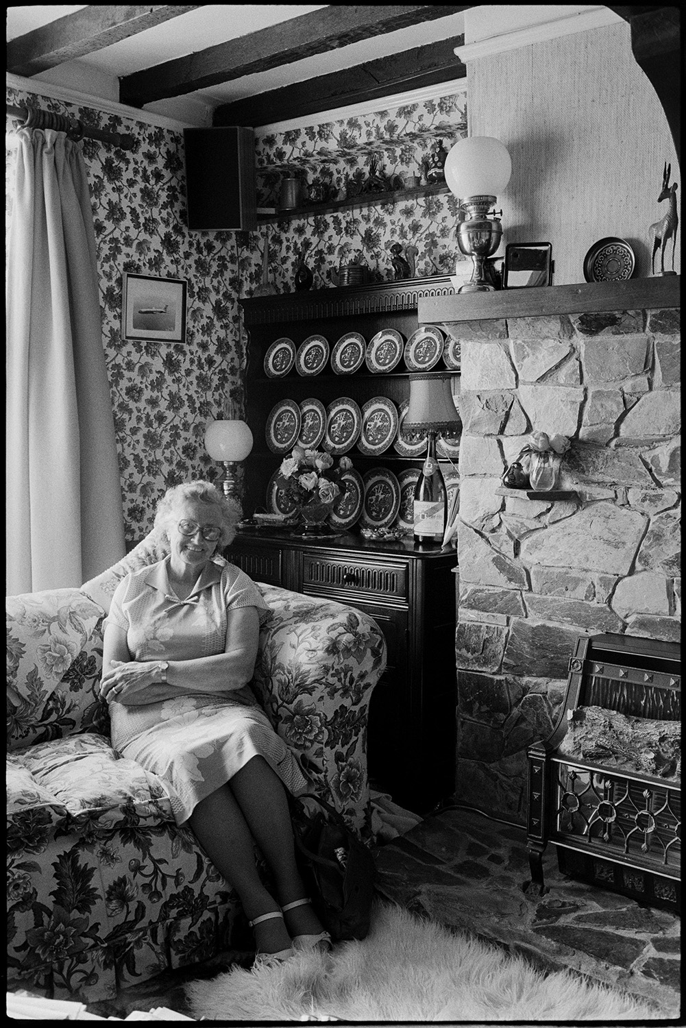 Woman seated in modernised olde worlde interior. Comparison with old photo.
[An interior shot of a room possibly in the Old Rectory, Weare Giffard, showing a woman sitting on a sofa in front of an oak cabinet containing a display of plates. The room has floral wallpaper and a stone fireplace, with an oil lamp on the mantelpiece. Timber beams are exposed in the ceiling.]