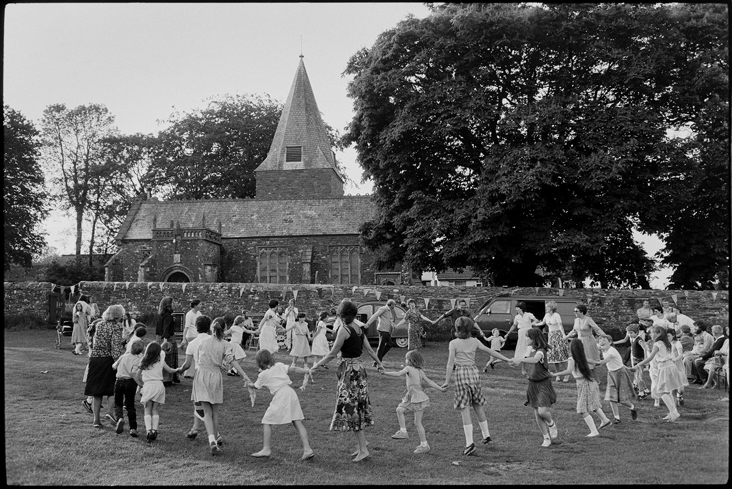 Children dancing on village green in front of church.
[Children, women and men holding hands in a circle and dancing on the Village Green at Beaford during the Beaford Revel. The Church of St George and All Saints is visible in the background, and a string of bunting is decorating the wall around the church.]