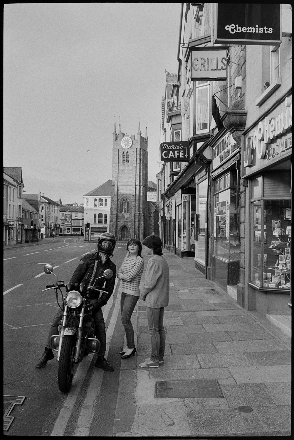 Street scenes, hotel and church tower.
[A young man seated on a motorbike talking to two young women in Fore Street, Okehampton, in front of a row of shops and cafes, including Maries' Café and a chemist. In the background the tower of St James' Church can be seen.]