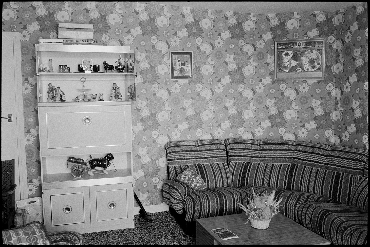 House interior, sitting room, pram and dresser, comparison with old photo, fitted kitchen. 
[Interior of a house in Lovacott Barton, Newton Tracey, showing a display of china ornaments on a dresser. Two pictures are hung on the wall which is decorated with floral wallpaper. A corner sofa and coffee table with a floral basket display are also visible.]