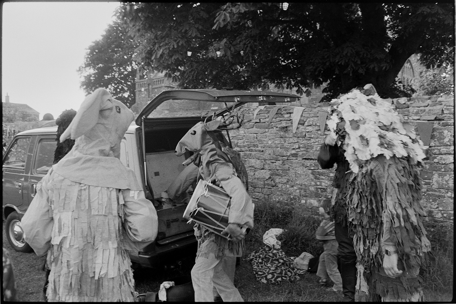 Morris men at revel.
[Three people dressed in costumes for a Mummers Play, one is holding a drum and unloading items from the back of a van parked next to the wall between the Church of St George and all Saints and Village Green in Beaford at the Beaford Revel.]