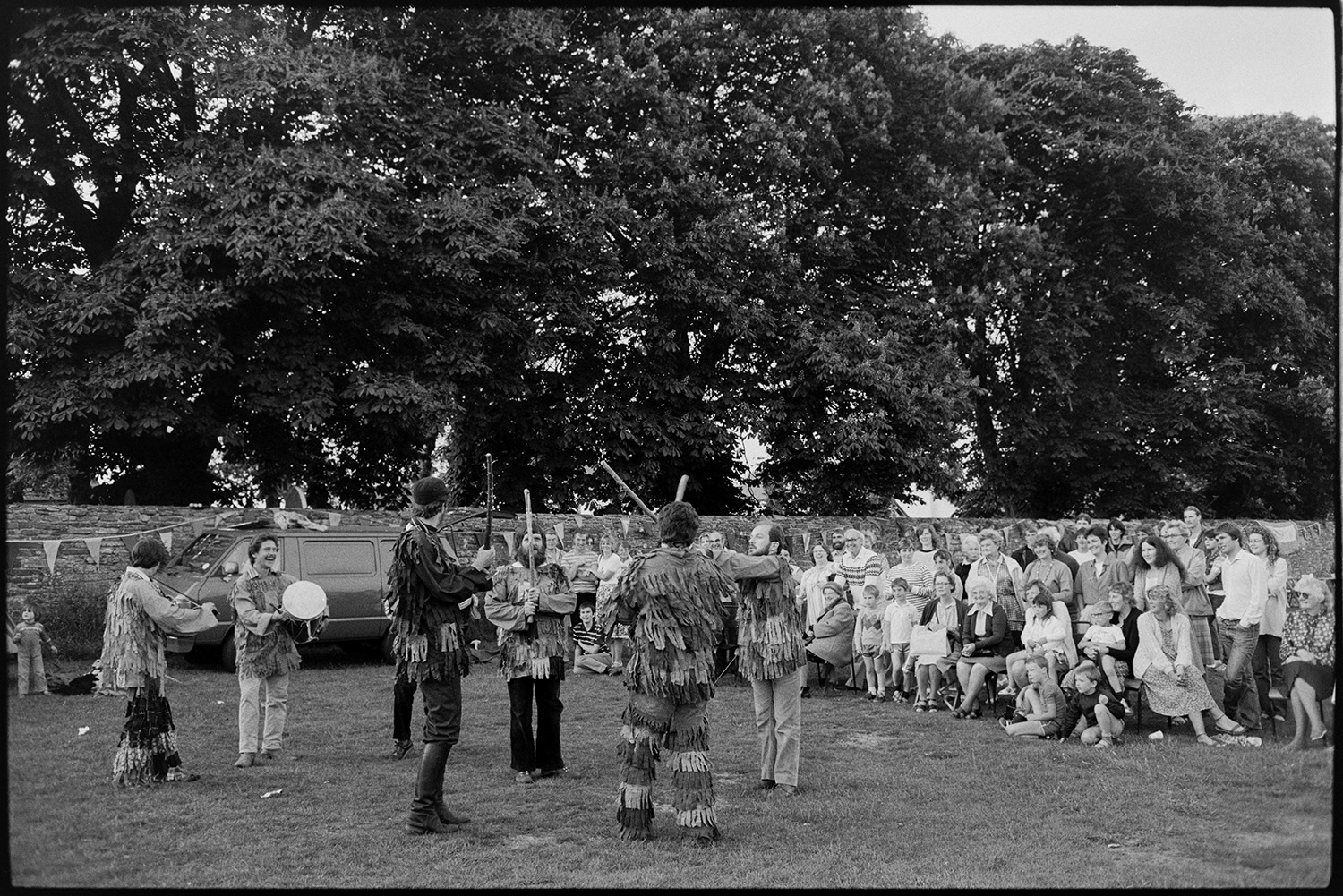 Morris men at revel.
[A group of Morris Men performing on the Village Green in Beaford at the Beaford Revel. Two of them are playing a drum and violin. They are being watched by a large group of men, women and children, some seated. A van is parked by a wall decorated with bunting in the background.]