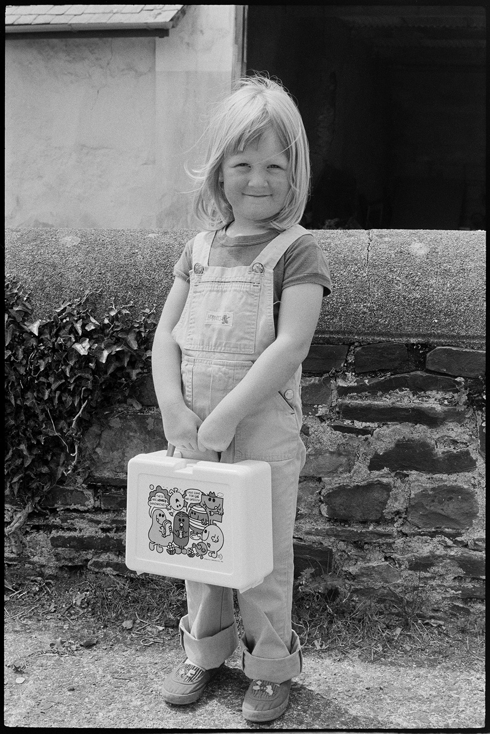 Little girl standing with lunch box.
[Sara Wright standing in a street in Dolton holding a plastic lunch box, decorated with Mr Men characters.]