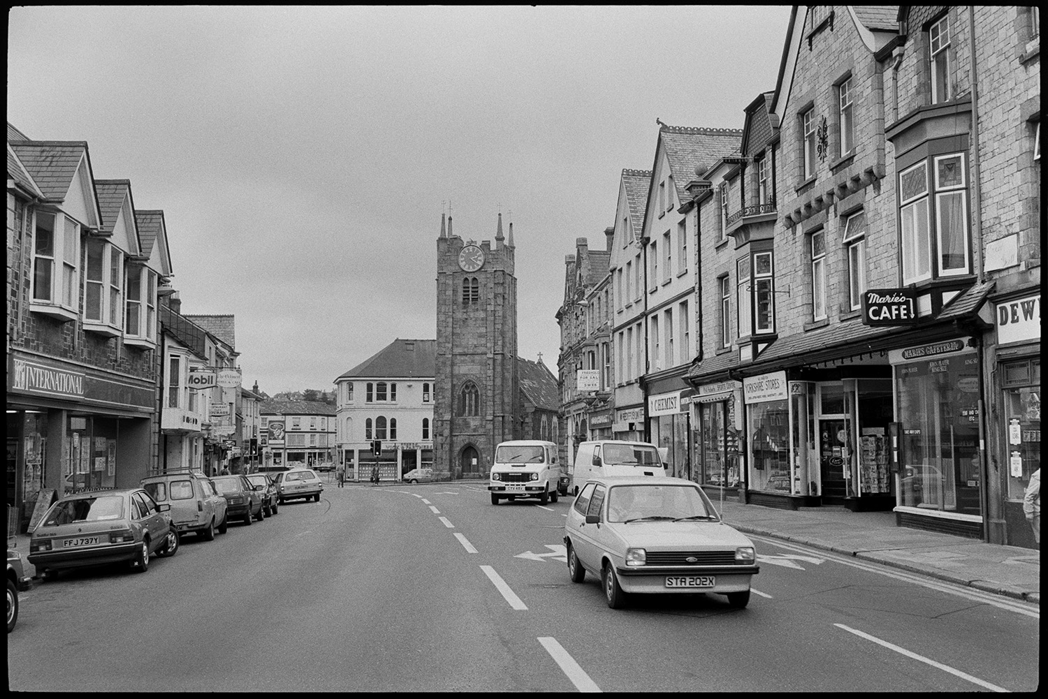 Street scene with cars, traffic church tower. Comparison with old photo.
[A view of Fore Street, Okehampton, showing St James' Church, and cars and vans parked and driving along the road. Shops including International Supermarket, Chemist, Yorkshire Stores, Maries' Cafe, Dewhurst, and Vick and Partners estate agents are visible.]