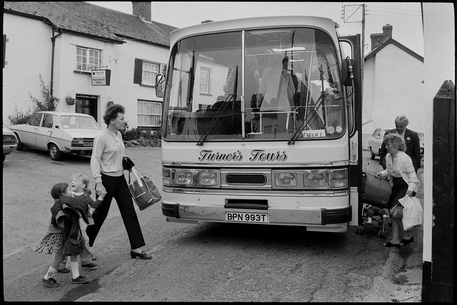 People getting onto bus.<br />
[Women, children and a man boarding a Turner's Tours coach to Chulmleigh in South Street, Dolton. In the background is a view of the Rams Head Inn and cars parked outside the Inn.]
